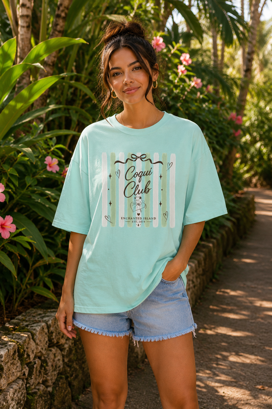 A woman wearing a light mint green graphic t-shirt with 'Coqui Club' printed on the front, paired with denim shorts, standing on a tropical garden path.