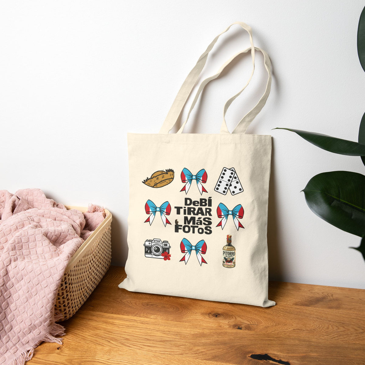 An off-white canvas tote bag with long handles stands on a wooden table against a white wall. The bag features a graphic print with the Spanish text 'DeBi TIRAR MIS FOTOS' surrounded by retro-style illustrations, including a camera, playing cards, a loaf of bread, and red-and-blue bows.