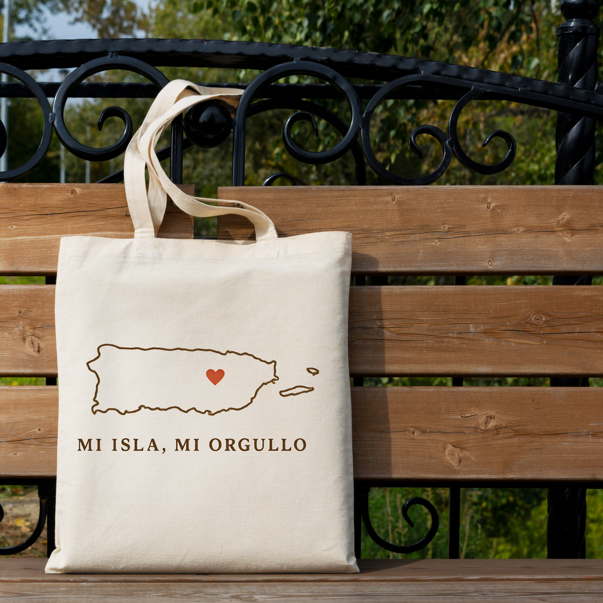 A natural canvas tote bag with a brown outline map of Puerto Rico, a small red heart, and the text 'MI ISLA, MI ORGULLO' resting on a wooden bench outdoors.