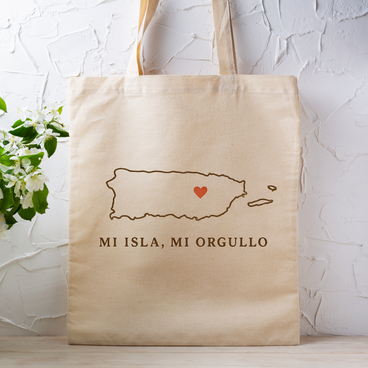 A beige canvas tote bag with a brown outline of Puerto Rico, a small red heart, and the text 'MI ISLA, MI ORGULLO' printed on the front, standing against a white textured wall with a small plant at the side.