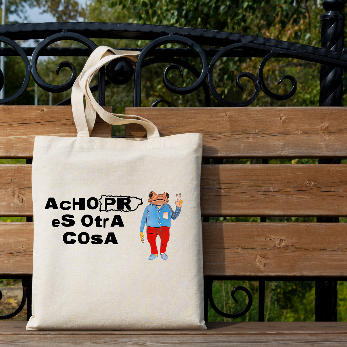 An off-white canvas tote bag with a humorous medical-themed graphic ('AChOPR eS OtrA COSA' and a cartoon figure in a blue coat and red pants) resting on a wooden park bench with a black metal railing and green foliage in the background.
