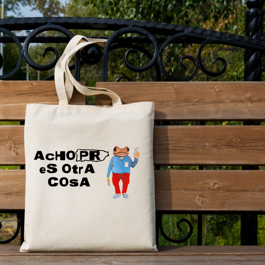 An off-white canvas tote bag with a humorous medical-themed graphic ('AChOPR eS OtrA COSA' and a cartoon figure in a blue coat and red pants) resting on a wooden park bench with a black metal railing and green foliage in the background.