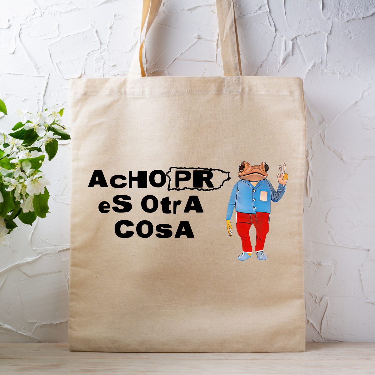 A beige canvas tote bag with a printed graphic of a frog in a blue shirt and red pants next to the text 'AchOPR eS OtrA CoSA', displayed against a white textured wall with a small plant on the left.