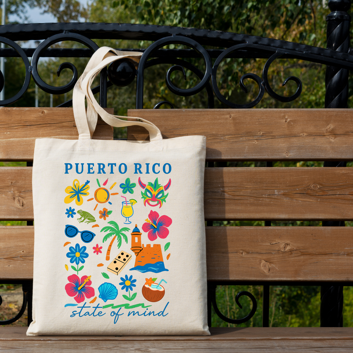 A cream canvas tote bag with 'PUERTO RICO' and colorful tropical illustrations hanging on a wooden bench outdoors. The bag features blue text and a multicolor print of flowers, palm trees, sunglasses, a beach umbrella, and a coconut.