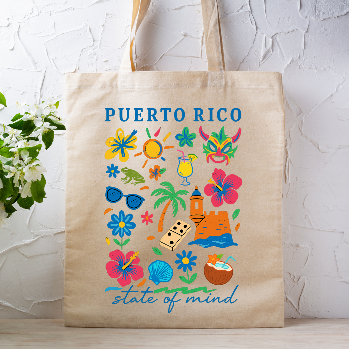 A beige canvas tote bag with a colorful Puerto Rico-themed graphic print, featuring tropical flowers, a mask, sunglasses, a drink, and the text 'PUERTO RICO' and 'state of mind', displayed against a white textured wall with a plant on the left.