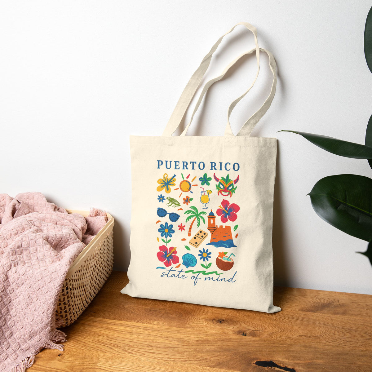 A cream canvas tote bag with 'PUERTO RICO' and 'state of mind' printed in blue, decorated with colorful tropical illustrations, leaning on a wooden table next to a pink blanket and a green plant.