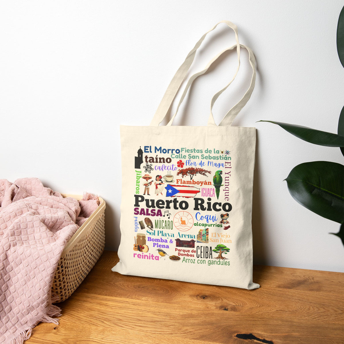 A cream canvas tote bag with colorful Puerto Rico-themed text and illustrations, standing on a wooden table next to a pink blanket and a green plant against a white wall.