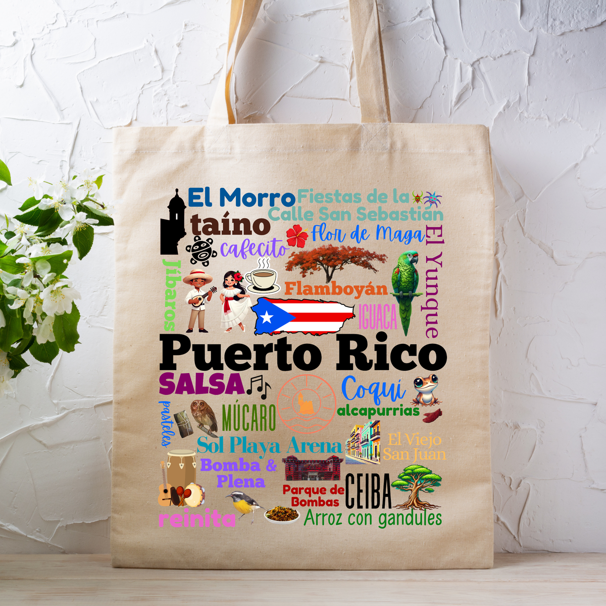 A beige canvas tote bag with a colorful Puerto Rico-themed collage print, displayed on a light wooden surface against a white textured wall with green leaves on the left.