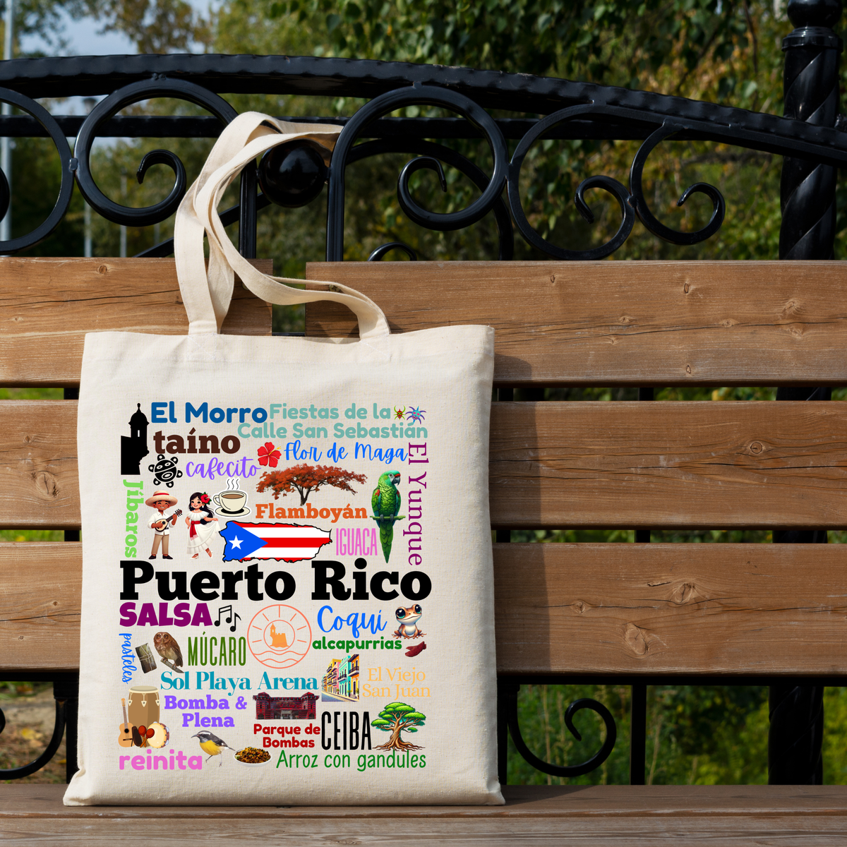 A cream canvas tote bag with 'Puerto Rico' and a colorful collage of cultural icons printed on the front, resting on a wooden bench with a black wrought iron railing and green foliage in the background.