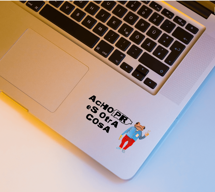 A close-up of a silver laptop showing the palm rest, keyboard, and trackpad, with a small printed sticker on the lower right of the palm rest. The sticker features the text 'ACHO PR eS OUA COSA' in black above a cartoon figure in a blue shirt and red pants.
