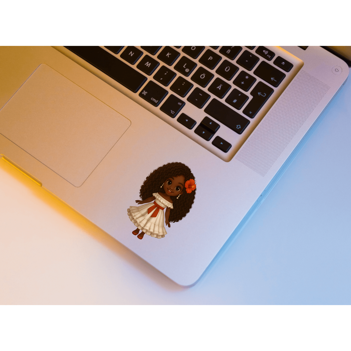 A die-cut vinyl sticker of a cartoon girl with dark skin, curly black hair, and a white dress with a red bow, shown applied to the palm rest of a silver laptop.
