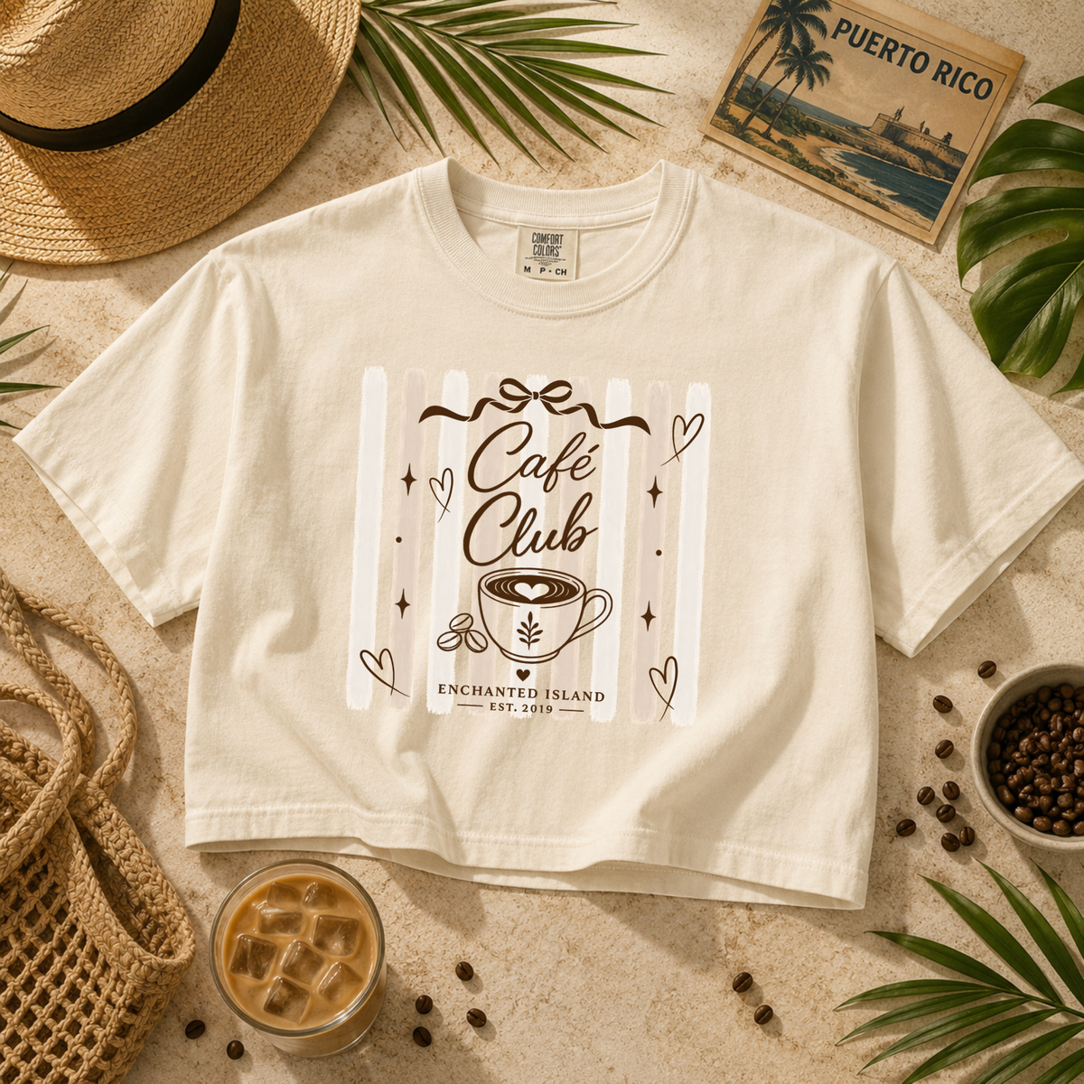 A cream-colored short-sleeve t-shirt with a brown graphic print reading 'Café Club Enchanted Island Est. 2019' and a coffee cup illustration, styled in a flat-lay with a straw hat, a woven bag, a glass of iced coffee, coffee beans, and a 'Puerto Rico' travel card.