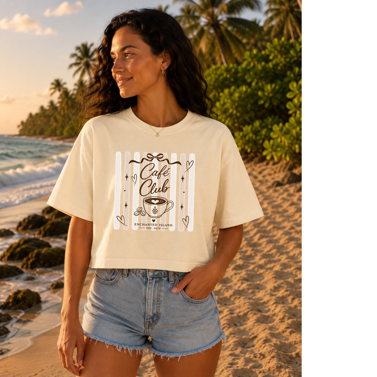 A woman stands on a sandy beach wearing a cream-colored graphic t-shirt with 'Cafe Club' written on it, paired with light-wash denim shorts.