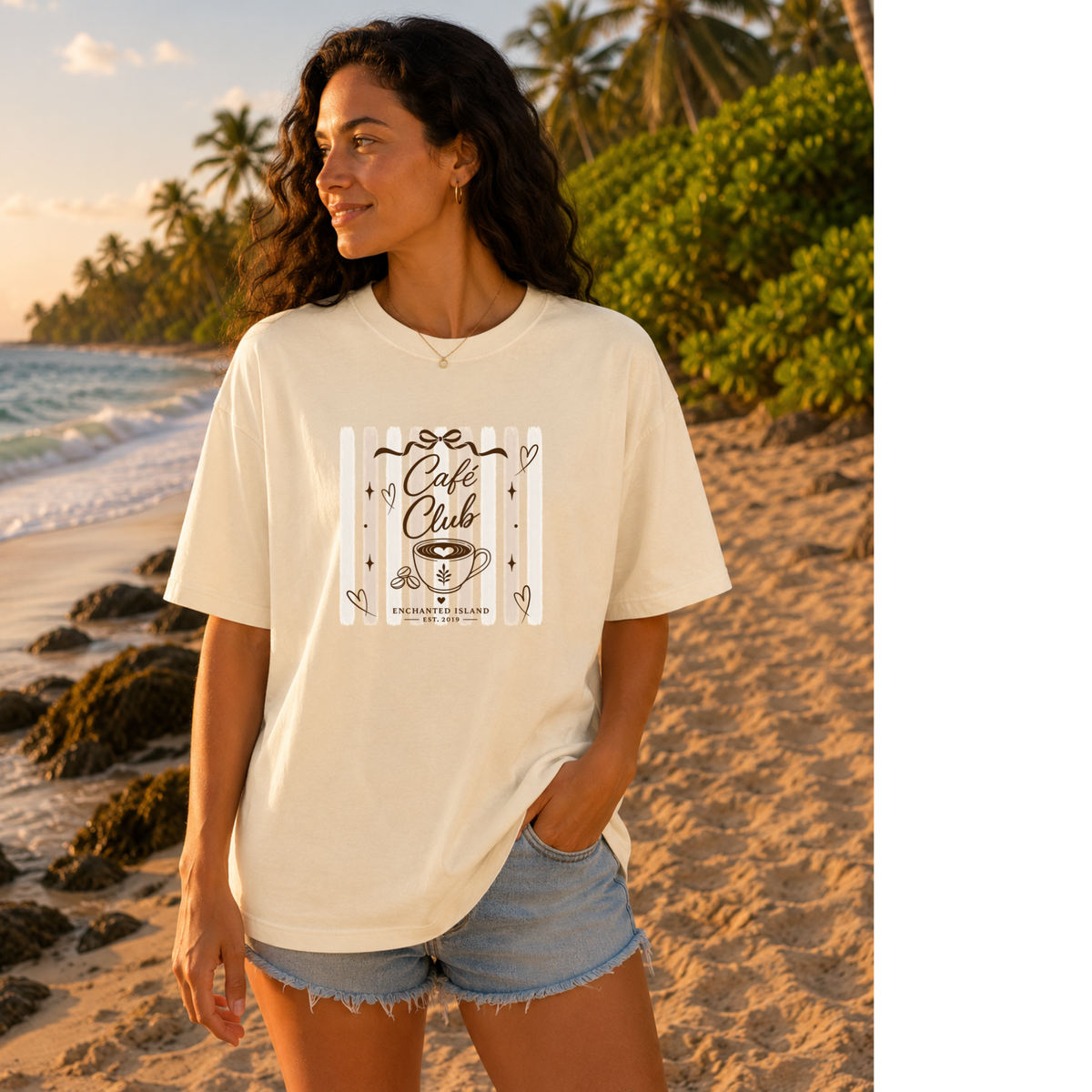 A woman stands on a sandy beach at sunset wearing an oversized cream-colored graphic T-shirt with 'Cafe Club' and a coffee cup illustration printed on the front, paired with denim shorts.