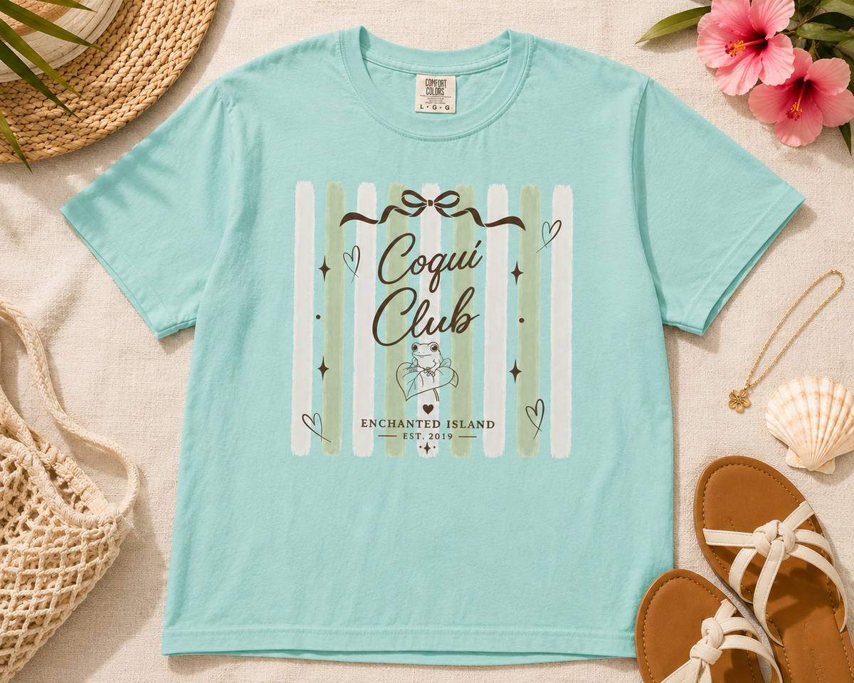 A light aqua short-sleeve graphic T-shirt laid flat on a beige fabric surface with beach-themed props including a straw hat, flowers, sandals, a woven bag, and a shell. The shirt has a central vertical stripe graphic in white and pale green with the words 'Coqui Club' and 'Enchanted Island' plus small decorative icons.