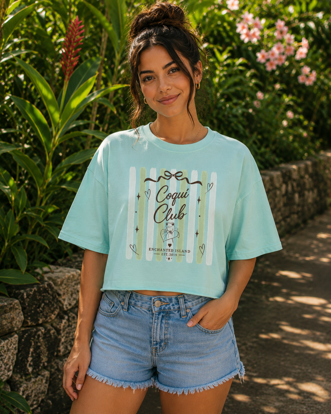 A woman wearing a light aqua blue graphic t-shirt and denim shorts, standing outdoors on a stone path surrounded by lush green foliage and pink flowers. The t-shirt features a graphic with the text 'Coqui Club' and 'BICHOLMEXICO'.