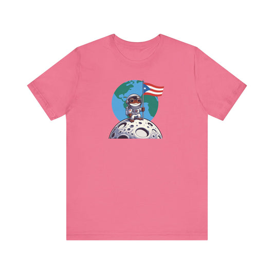 A pink short-sleeve crewneck t-shirt featuring a cartoon graphic of an astronaut standing on a cratered moon with a blue globe and a red, white, and blue flag in the background.