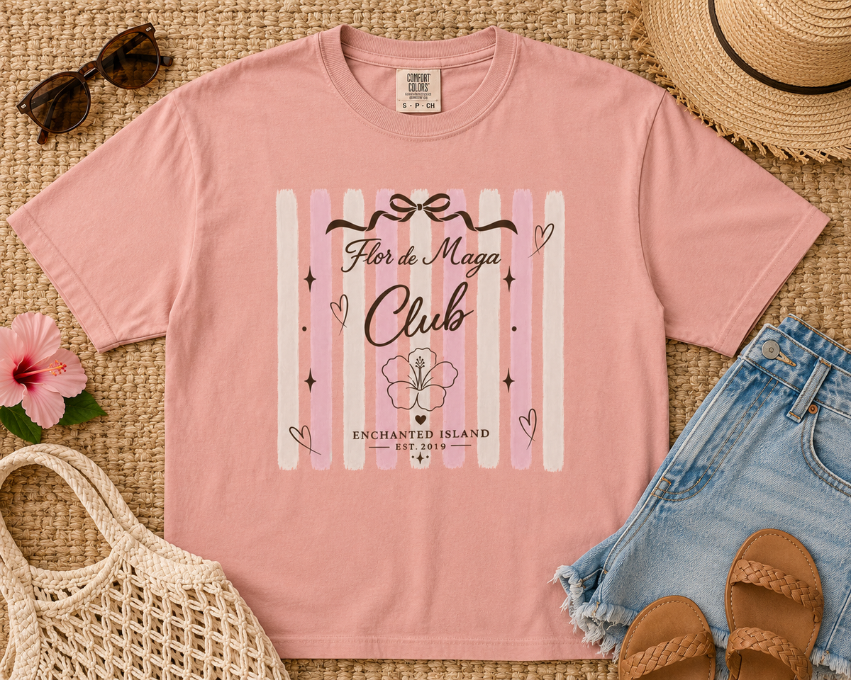 A pink graphic t-shirt laid flat on a woven straw mat, surrounded by summer accessories including denim shorts, a straw hat, sunglasses, a woven bag, and sandals. The t-shirt features a vertical striped design in shades of pink and white with the text 'Flor de Maga Club' and 'Enchanted Island Est.