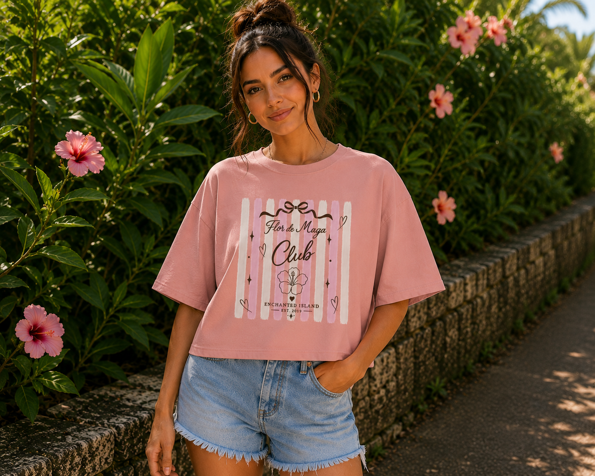 A woman wearing a pink graphic t-shirt and denim shorts stands outdoors on a paved path surrounded by green foliage and pink flowers. The t-shirt features a white and pink striped graphic with the text 'Flower Mfg Club' and 'Palm Beaches, FL'.
