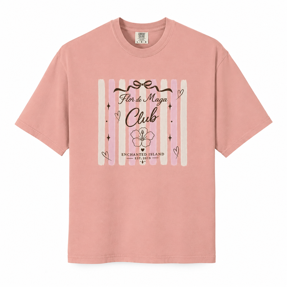A dusty pink short-sleeve t-shirt with a graphic on the front featuring vertical pastel stripes, a flower illustration, and the text 'Flor de Magia Club' and 'Enchanted Island'.