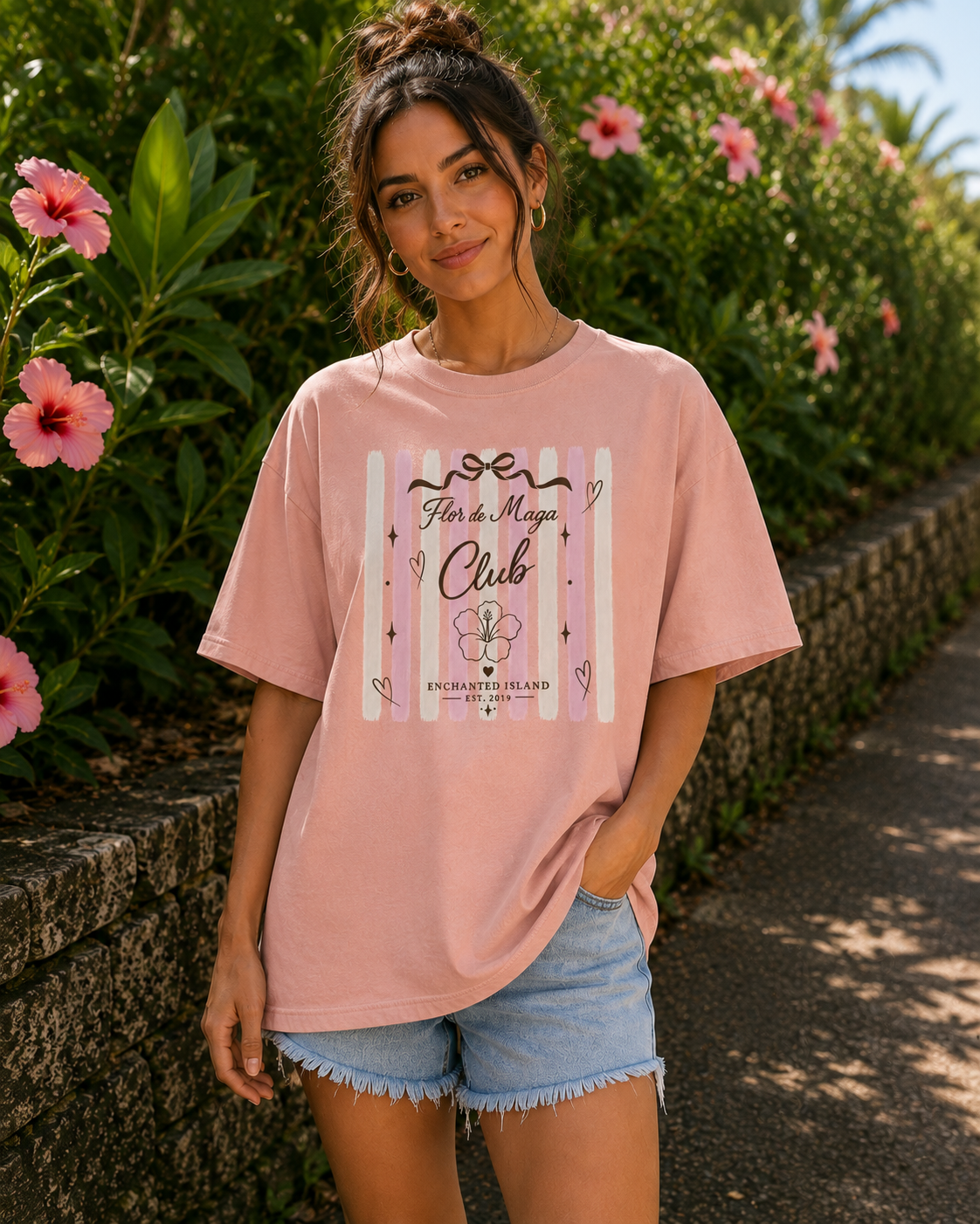 A woman wearing a light pink graphic t-shirt and denim shorts stands outdoors on a paved path with green foliage and pink flowers in the background. The t-shirt features a graphic design with the text 'Fun to Mega Club' and 'FUKUSHIMARUO' on a striped background.