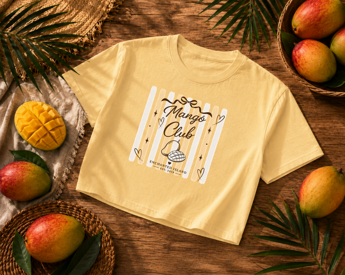 A pale yellow short-sleeve crop top T-shirt laid flat on a wooden surface, surrounded by mangoes, palm leaves, and woven baskets. The shirt has a large front graphic with the words 'Mango Club' over white vertical bars and a small illustration below.