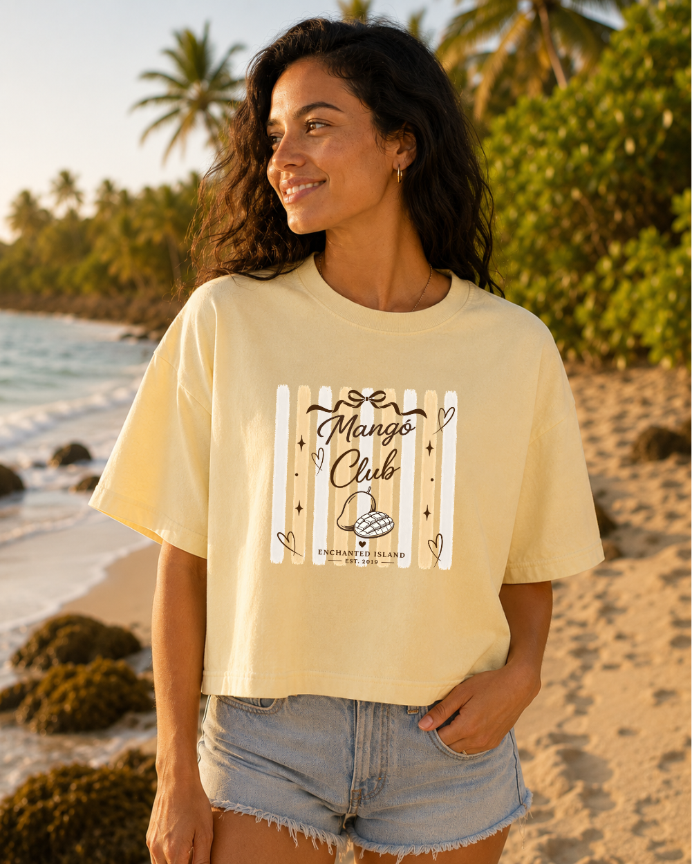 A woman stands on a sandy beach wearing a pale yellow graphic T-shirt with 'Mango Club' and 'King of the Hill' artwork, paired with light denim shorts. Palm trees and ocean are visible in the background.