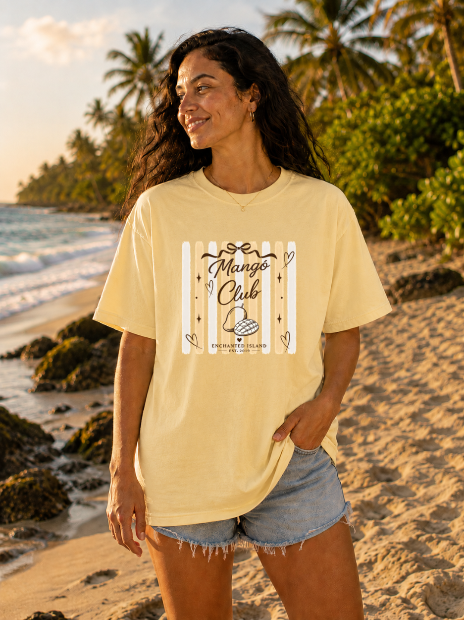 A woman stands on a sandy beach wearing a pale yellow graphic T-shirt that reads 'Mango Club' with a mango illustration and 'Pura Beloved Islands' below, paired with denim shorts. Palm trees and the ocean are visible in the background.